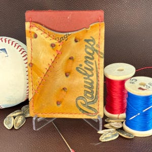 May include: A handmade cardholder crafted from baseball glove leather, featuring the Rawlings logo. The cardholder is displayed with a baseball, spools of red and blue thread, and a needle. The cardholder is tan with red stitching.