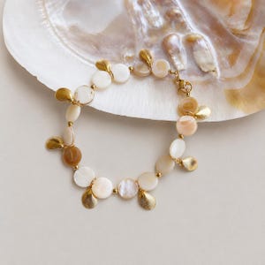 May include: A bracelet featuring circular white and tan shell beads interspersed with small gold-toned metal beads and dangling gold-toned metal shell charms. The bracelet is arranged in a circular shape and includes a gold-toned metal lobster clasp.