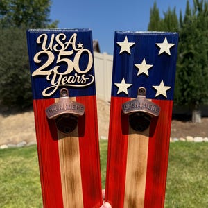 May include: Two rectangular wooden wall-mounted bottle openers painted with red, white, and blue stripes. The left piece features the text USA 250 Years in raised lettering. The right piece displays five white stars. Both have a bronze-toned metal opener.