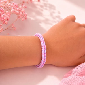 May include: A braided bracelet in pink and lavender hues is worn on a wrist. The bracelet is made of small, interwoven bands. The background features pink flowers and a pink cloth.