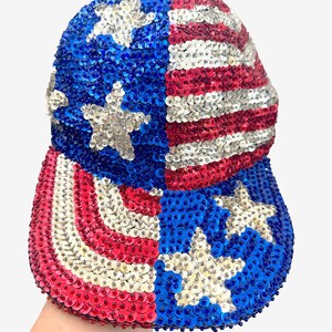 May include: A baseball cap completely covered in shimmering sequins arranged in a pattern resembling the American flag. The design features blue sections with white stars and alternating red and white stripes across the crown and the curved brim.