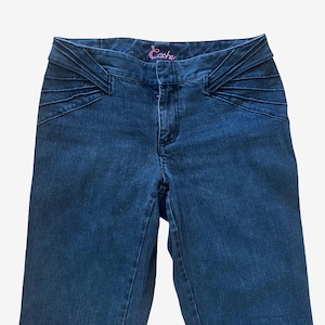 May include: Dark blue denim jeans featuring a wide waistband with decorative horizontal pleats on both sides. The interior waistband label displays the word Cache in pink cursive embroidery. The jeans have a standard button and zipper fly closure.