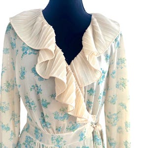 May include: A long-sleeved wrap dress in an off-white color featuring a small blue floral print throughout. The garment has a prominent, wide, pleated cream-colored ruffle trim that extends along the V-neckline and down the front bodice.