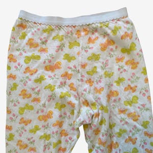 May include: White leggings featuring an all-over print of orange and lime green butterflies interspersed with small pink flower clusters. The garment has a white elastic waistband with a subtle scalloped edge detail along the top seam.