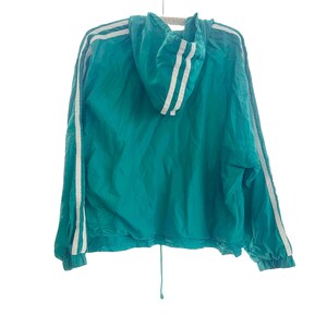 May include: A teal windbreaker jacket featuring a hood and long sleeves with two white stripes running down each arm. The jacket has elastic cuffs at the wrists and a drawstring cord at the bottom hem, providing a cinched fit at the waistline.