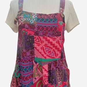 May include: A sleeveless pinafore dress featuring a vibrant patchwork design with various geometric and paisley patterns in shades of pink, purple, blue, and green, worn over a short-sleeved white ribbed t-shirt with a round neckline.