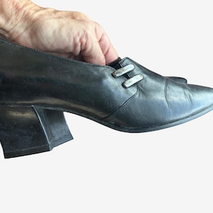 May include: A black leather shoe with a pointed toe, a thick block heel, and two silver-toned metal rectangular accents on the side near the opening. The shoe features a smooth finish and a structured silhouette with a low-cut vamp design.
