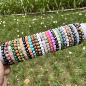 Peut inclure: Une collection de nombreux bracelets extensibles composés de diverses perles en pierre naturelle colorées, incluant des tons de vert, rose, bleu, marron, noir et blanc, disposés côte à côte sur un présentoir cylindrique gris.