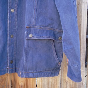 May include: A blue denim jacket with a button-up front, two flap pockets, and a single pocket on the left sleeve. The jacket has a corduroy collar and a brown stitching detail.