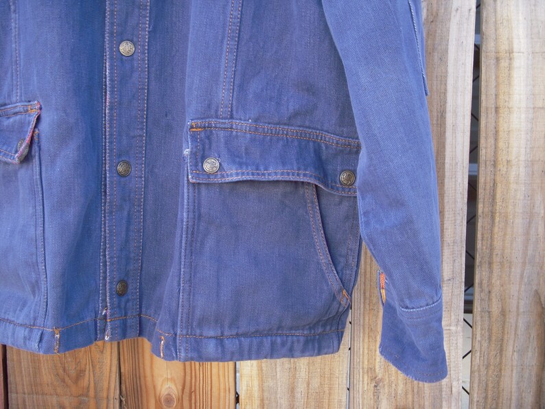 May include: A blue denim jacket with a button-up front, two flap pockets, and a single pocket on the left sleeve. The jacket has a corduroy collar and a brown stitching detail.
