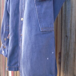 May include: A blue denim jacket with a button-up front and a patch pocket on the right sleeve. The jacket has a faded look and some white marks on the sleeve.