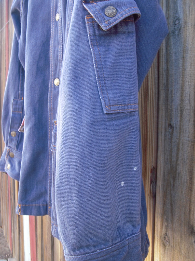 May include: A blue denim jacket with a button-up front and a patch pocket on the right sleeve. The jacket has a faded look and some white marks on the sleeve.