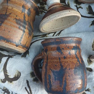 May include: Handmade ceramic mugs and goblets in shades of brown and blue. The pottery features a rustic, textured finish with unique glaze patterns. The mugs have handles, and the goblets have a stem and a wide base. The items are arranged on a floral patterned cloth.