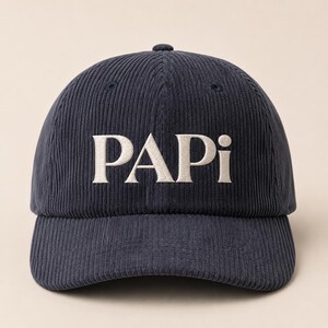 May include: A dark navy blue corduroy baseball cap featuring the word PAPI embroidered in white capital letters across the front panel. The hat has a structured crown, a curved brim, and a button at the top centre of the fabric dome.