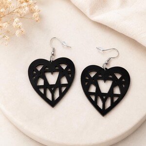 May include: A pair of black heart-shaped earrings with a geometric design. The earrings have a cutout star design in the centre and are suspended from silver-coloured fish hook ear wires. The earrings are displayed on a white surface.