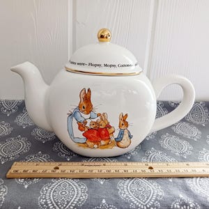 May include: A white ceramic teapot with a gold-tone knob on the lid and a Peter Rabbit illustration featuring four rabbits. The text on the lid reads, "their names were-- Flopsy, Mopsy, Cotton-tail and Peter." A wooden ruler shows the teapot is 10 inches wide.