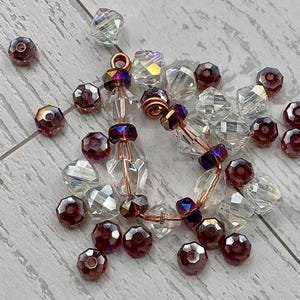 May include: A collection of faceted beads in clear and deep burgundy colors, some strung on copper wire with a small loop at the top. The beads vary in shape, including round, bicone, and elongated forms, with iridescent finishes on the clear pieces.