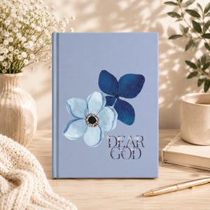 Puede incluir: Un diario de tapa dura color azul claro con una ilustración en acuarela de una flor azul claro y una flor azul oscuro. El texto "DEAR GOD" está impreso en una fuente serif azul oscuro debajo del diseño floral en la portada.