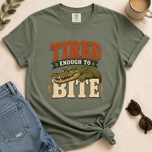 May include: An olive green crew neck t-shirt featuring a graphic of a crocodile with the text TIRED ENOUGH TO BITE in orange and cream lettering. The design includes small yellow lightning bolt accents and a dark teal banner element.