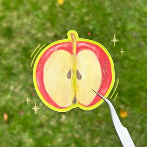 Puede incluir: Una pegatina troquelada que muestra un corte transversal de una manzana roja con interior amarillo pálido y semillas marrones visibles. La pegatina tiene un borde amarillo brillante y es sostenida por pinzas de metal plateado sobre fondo verde.