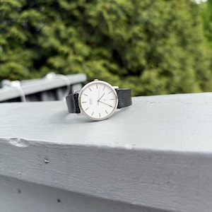 May include: A silver-tone wristwatch with a round white dial, thin black hour markers, and black hands. The watch features a textured black leather strap and the text Seiko Quartz printed on the face. The design includes a small heart icon.