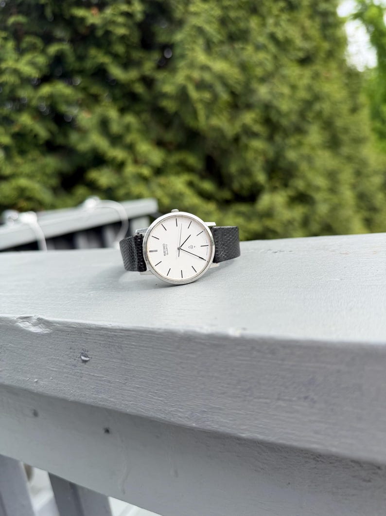 May include: A silver-tone wristwatch with a round white dial, thin black hour markers, and black hands. The watch features a textured black leather strap and the text Seiko Quartz printed on the face. The design includes a small heart icon.