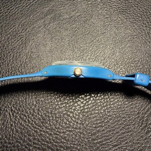 K&ouml;nnte beinhalten: Eine leuchtend blaue Armbanduhr in Seitenansicht. Die Uhr verf&uuml;gt &uuml;ber ein glattes, mattblaues Kunststoffgeh&auml;use und ein passendes blaues Armband. Eine kleine, kreisf&ouml;rmige, silberfarbene Krone befindet sich an der Seite des Geh&auml;uses zur Zeiteinstellung.