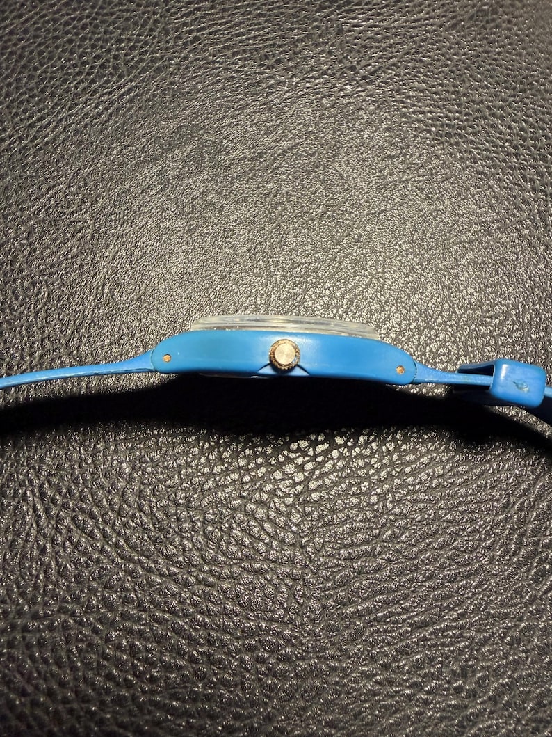 K&ouml;nnte beinhalten: Eine leuchtend blaue Armbanduhr in Seitenansicht. Die Uhr verf&uuml;gt &uuml;ber ein glattes, mattblaues Kunststoffgeh&auml;use und ein passendes blaues Armband. Eine kleine, kreisf&ouml;rmige, silberfarbene Krone befindet sich an der Seite des Geh&auml;uses zur Zeiteinstellung.