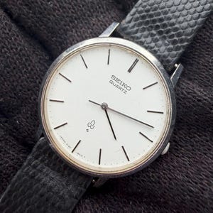 May include: A Seiko Quartz wristwatch with a round silver-tone case and a white dial featuring thin black hour markers and hands. The watch includes a black textured leather strap and displays the Seiko Quartz logo and a small emblem on the face.