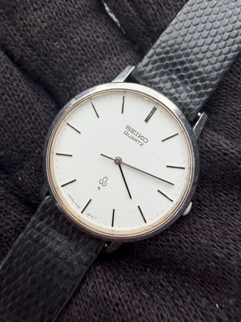 May include: A Seiko Quartz wristwatch with a round silver-tone case and a white dial featuring thin black hour markers and hands. The watch includes a black textured leather strap and displays the Seiko Quartz logo and a small emblem on the face.