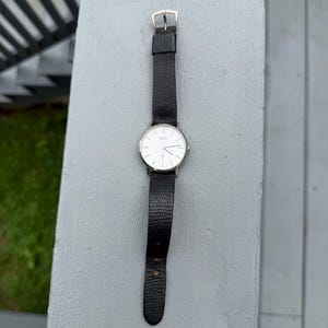 May include: A Seiko quartz wristwatch featuring a round silver-tone case with a white dial, thin black hour markers, and slender hands. The watch is attached to a black leather strap with a textured, lizard-skin pattern and a silver-tone buckle.