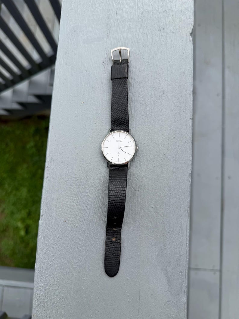 May include: A Seiko quartz wristwatch featuring a round silver-tone case with a white dial, thin black hour markers, and slender hands. The watch is attached to a black leather strap with a textured, lizard-skin pattern and a silver-tone buckle.