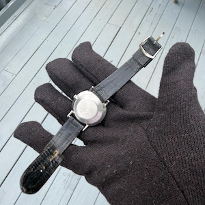 May include: A wristwatch with a round silver-tone metal case and a textured black leather strap. The back of the watch case is circular and metallic, featuring a central circular indentation. The strap includes a silver-tone metal buckle.