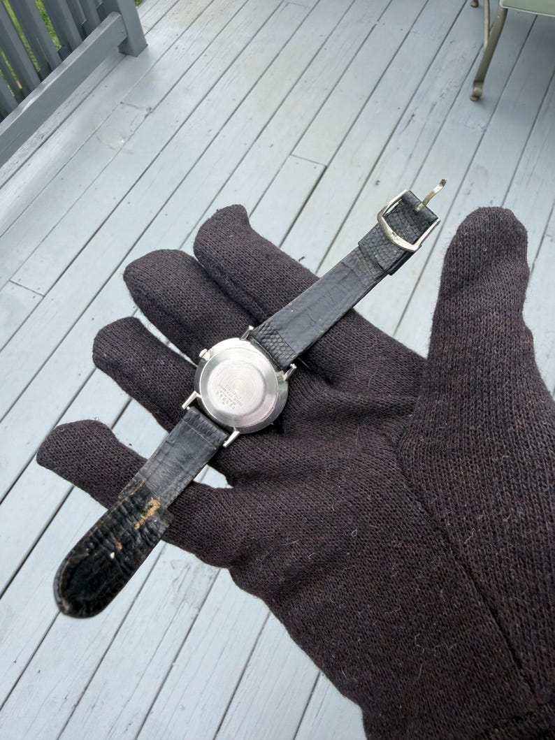 May include: A wristwatch with a round silver-tone metal case and a textured black leather strap. The back of the watch case is circular and metallic, featuring a central circular indentation. The strap includes a silver-tone metal buckle.