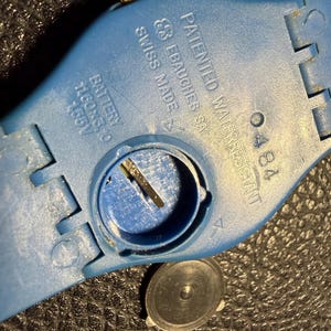 K&ouml;nnte beinhalten: Ein blaues Kunststoff-Uhrengeh&auml;use mit einem runden Batteriefach und einem abgenommenen Deckel. Der Text lautet: PATENTED WATERRESISTANT, EBAUCHES SA, SWISS MADE, 0484 und BATTERY 11.60x3.10 1.55V. Eine kleine metallische Krone ist am Rand zu sehen.