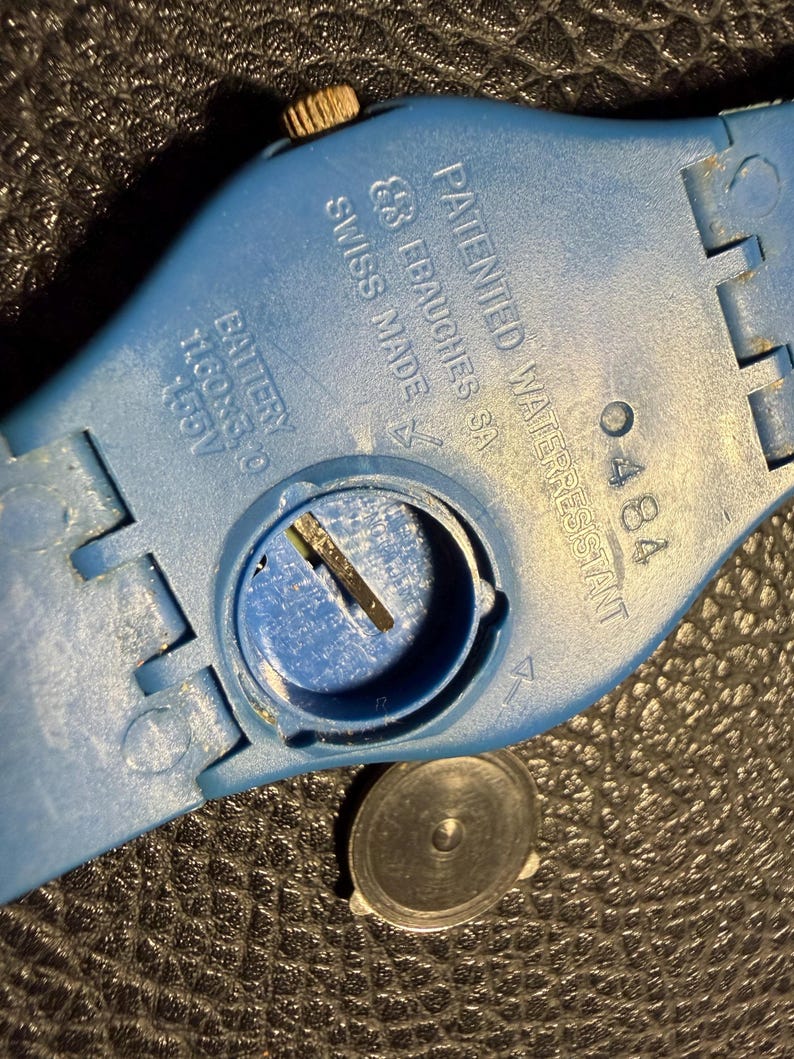 K&ouml;nnte beinhalten: Ein blaues Kunststoff-Uhrengeh&auml;use mit einem runden Batteriefach und einem abgenommenen Deckel. Der Text lautet: PATENTED WATERRESISTANT, EBAUCHES SA, SWISS MADE, 0484 und BATTERY 11.60x3.10 1.55V. Eine kleine metallische Krone ist am Rand zu sehen.