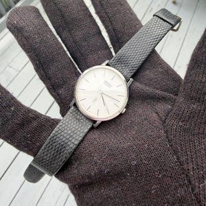 May include: A silver-tone Seiko Quartz wristwatch with a round white dial, thin silver hour markers, and slender hands. The watch features a black textured strap with a small metal buckle, showcasing a clean and simple design for daily timekeeping.