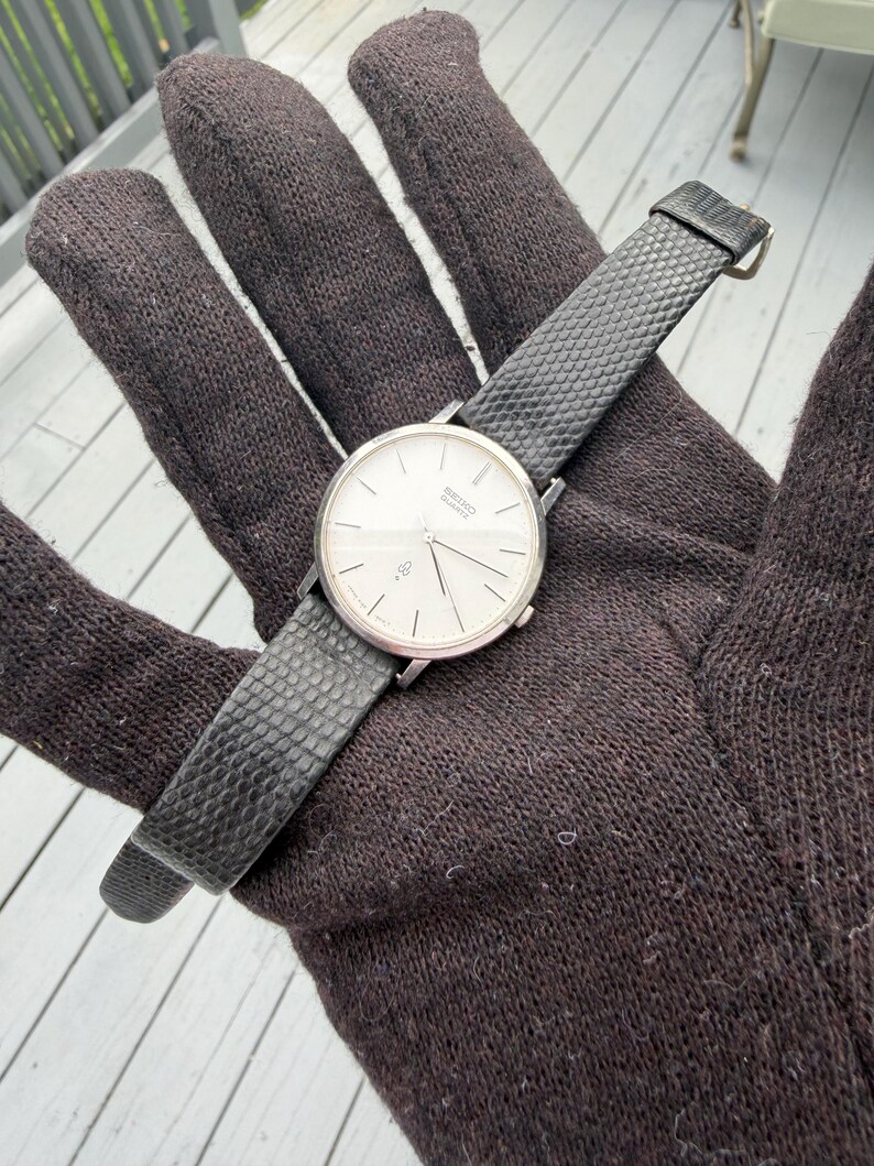 May include: A silver-tone Seiko Quartz wristwatch with a round white dial, thin silver hour markers, and slender hands. The watch features a black textured strap with a small metal buckle, showcasing a clean and simple design for daily timekeeping.