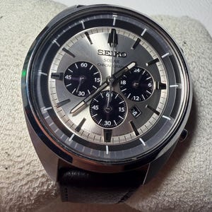 Puede incluir: Un reloj cronógrafo Seiko Solar con esfera plateada efecto rayos de sol, tres subesferas negras y correa de cuero negro. La esfera presenta índices plateados, manecillas luminiscentes y una ventana de fecha en la posición de las cuatro.