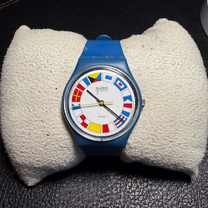 K&ouml;nnte beinhalten: Eine blaue Swatch Quartz Armbanduhr mit wei&szlig;em Zifferblatt, das von bunten nautischen Signalflaggen umrandet ist. Die Uhr verf&uuml;gt &uuml;ber ein blaues Silikonarmband, eine silberfarbene Krone an der rechten Seite sowie schwarze Zeiger.