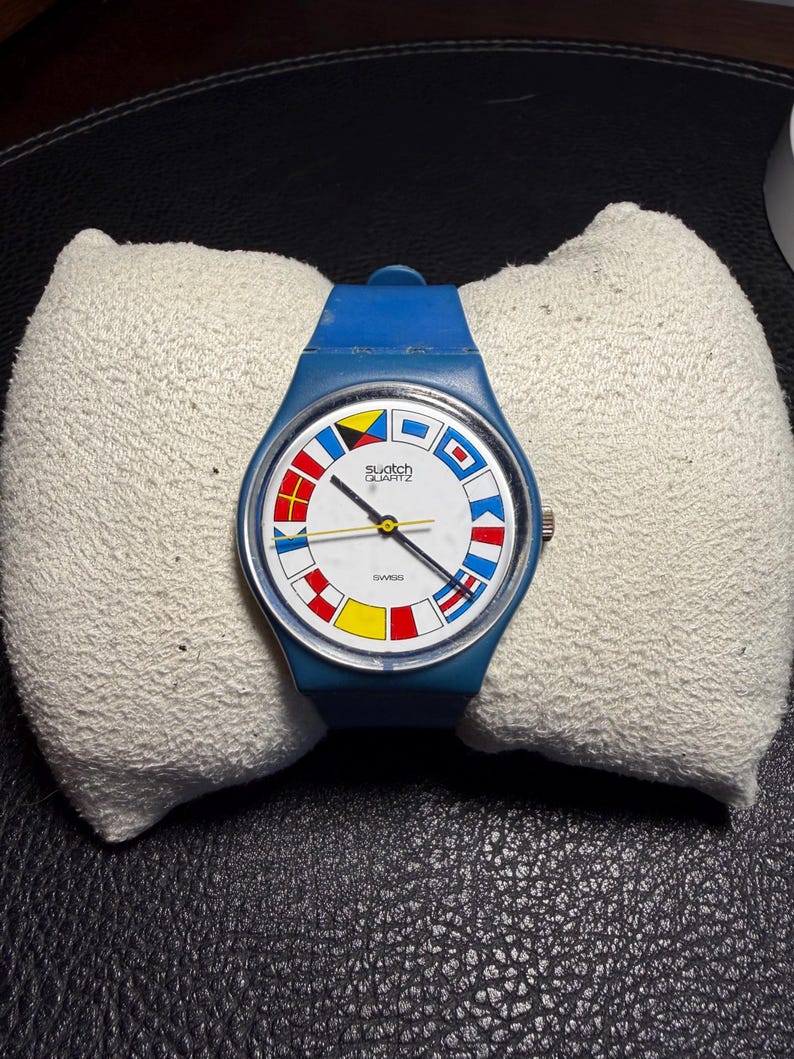 K&ouml;nnte beinhalten: Eine blaue Swatch Quartz Armbanduhr mit wei&szlig;em Zifferblatt, das von bunten nautischen Signalflaggen umrandet ist. Die Uhr verf&uuml;gt &uuml;ber ein blaues Silikonarmband, eine silberfarbene Krone an der rechten Seite sowie schwarze Zeiger.
