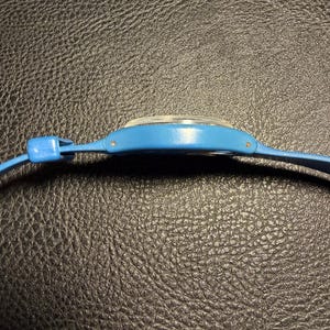 K&ouml;nnte beinhalten: Eine Seitenansicht einer leuchtend blauen Armbanduhr mit einem glatten, geschwungenen Kunststoffgeh&auml;use und einem passenden blauen Armband. Die Uhr hat ein klares, leicht gew&ouml;lbtes Kristallglas und kleine Metallstifte zur Befestigung des Armbands.