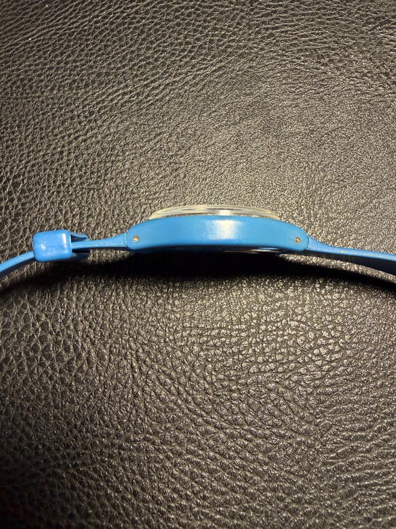 K&ouml;nnte beinhalten: Eine Seitenansicht einer leuchtend blauen Armbanduhr mit einem glatten, geschwungenen Kunststoffgeh&auml;use und einem passenden blauen Armband. Die Uhr hat ein klares, leicht gew&ouml;lbtes Kristallglas und kleine Metallstifte zur Befestigung des Armbands.