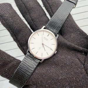 May include: A Seiko Quartz wristwatch featuring a round silver-tone case, a white dial with thin black hour markers, and a textured black strap. The watch face displays the Seiko Quartz logo and a small symbol near the six o'clock position.