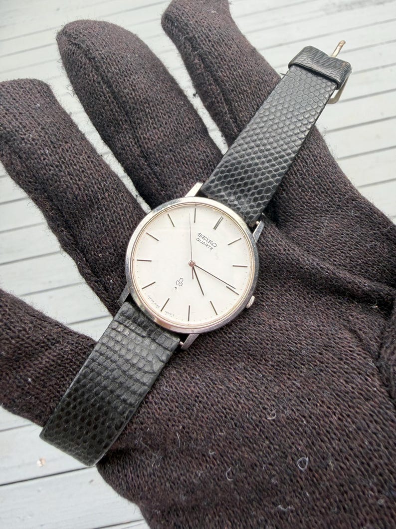 May include: A Seiko Quartz wristwatch featuring a round silver-tone case, a white dial with thin black hour markers, and a textured black strap. The watch face displays the Seiko Quartz logo and a small symbol near the six o'clock position.