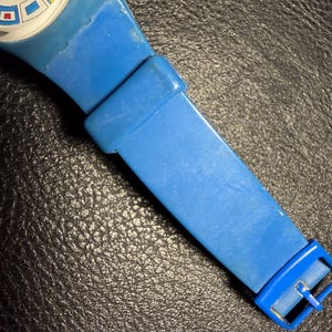 K&ouml;nnte beinhalten: Eine blaue Swatch Quarzuhr mit einem wei&szlig;en Zifferblatt und bunten geometrischen Markierungen. Die Uhr verf&uuml;gt &uuml;ber ein einfarbiges blaues Kunststoffarmband mit passender blauer Schlie&szlig;e und Schlaufe. Der Markenname Swatch steht auf dem Zifferblatt.