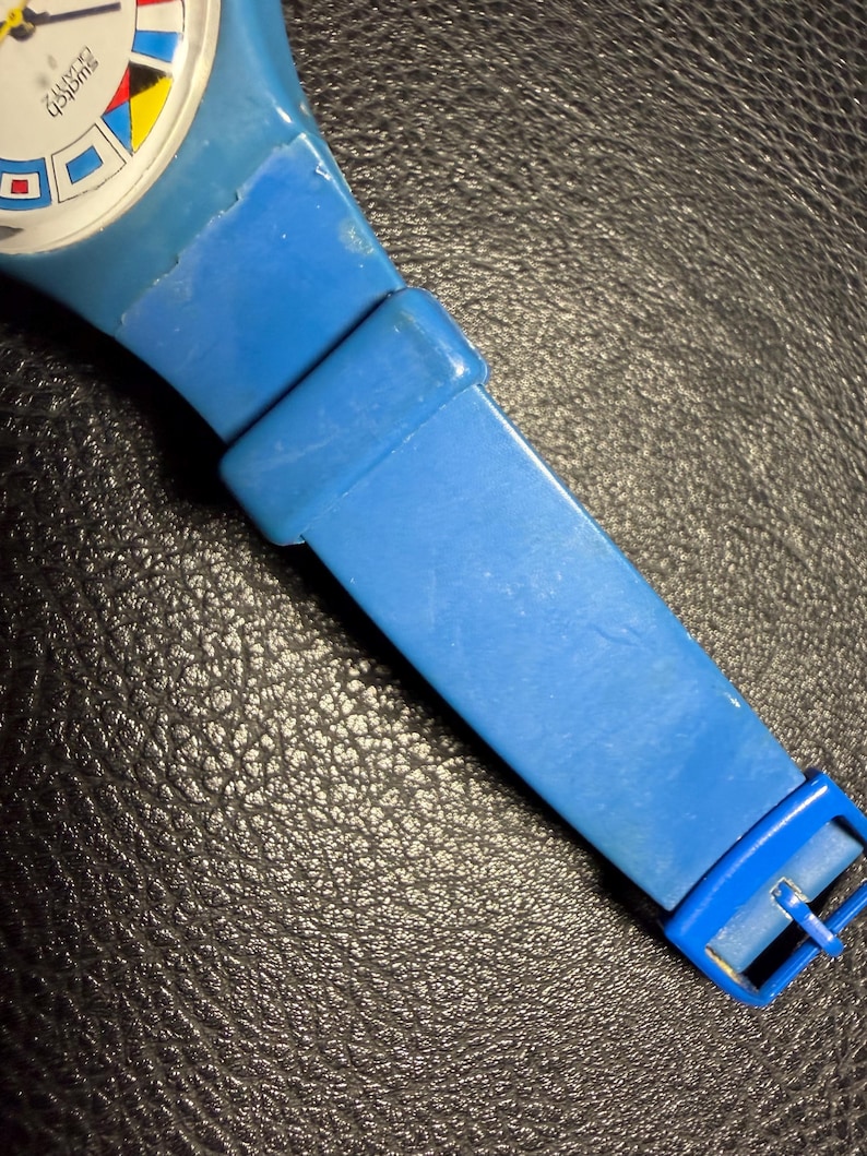 K&ouml;nnte beinhalten: Eine blaue Swatch Quarzuhr mit einem wei&szlig;en Zifferblatt und bunten geometrischen Markierungen. Die Uhr verf&uuml;gt &uuml;ber ein einfarbiges blaues Kunststoffarmband mit passender blauer Schlie&szlig;e und Schlaufe. Der Markenname Swatch steht auf dem Zifferblatt.