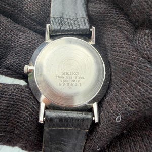 May include: A stainless steel Seiko watch case back with a black leather strap. The metal back features the engraved text SEIKO, STAINLESS STEEL, 4120-9010, and the serial number 690531. The circular metal surface has a brushed, radial finish.