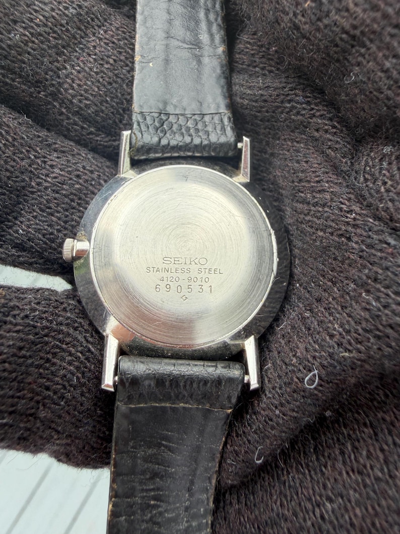 May include: A stainless steel Seiko watch case back with a black leather strap. The metal back features the engraved text SEIKO, STAINLESS STEEL, 4120-9010, and the serial number 690531. The circular metal surface has a brushed, radial finish.