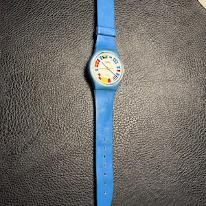 K&ouml;nnte beinhalten: Eine blaue analoge Armbanduhr mit einem runden wei&szlig;en Zifferblatt, das bunte nautische Signalflaggen als Stundenmarkierungen zeigt. Die Uhr hat ein passendes blaues Silikonarmband mit Dornschlie&szlig;e sowie goldfarbene Zeiger.